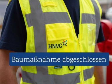 Mitarbeitende Person in gelber Warnweste mit HNVG-Logo in industrieller Umgebung.