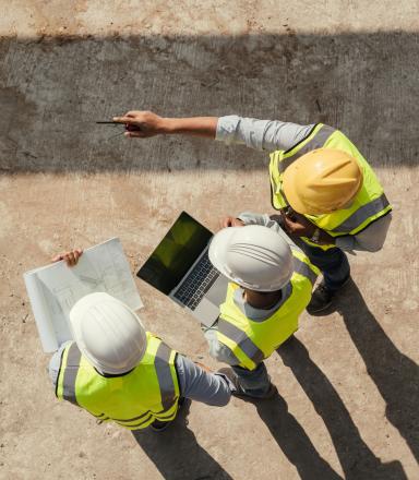 Baustellenbesprechung: Drei Bauingenieure mit Helm und Warnweste planen ein Projekt mit Laptop und Bauplänen