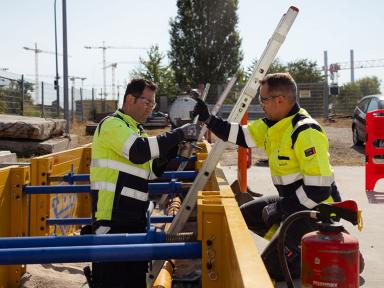 Zwei Bauarbeiter in Warnschutzkleidung montieren auf einer Baustelle eine Leiter an einer gelben Baugrubenabsperrung.