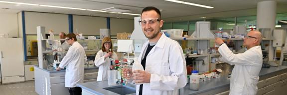 Ein Mann in einem Laborkittel hält einen Behälter und eine Flasche.