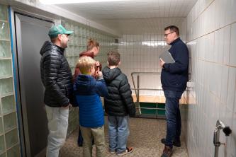 Familie mit zwei Kindern erhält in einem schmalen, gefliesten Kellerraum eine Führung durch einen erklärenden Mitarbeiter.