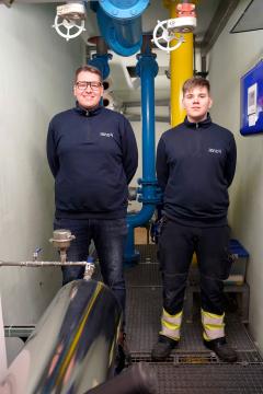 Zwei Personen in dunkelblauer Arbeitskleidung stehen in einem Betriebstechnikraum.